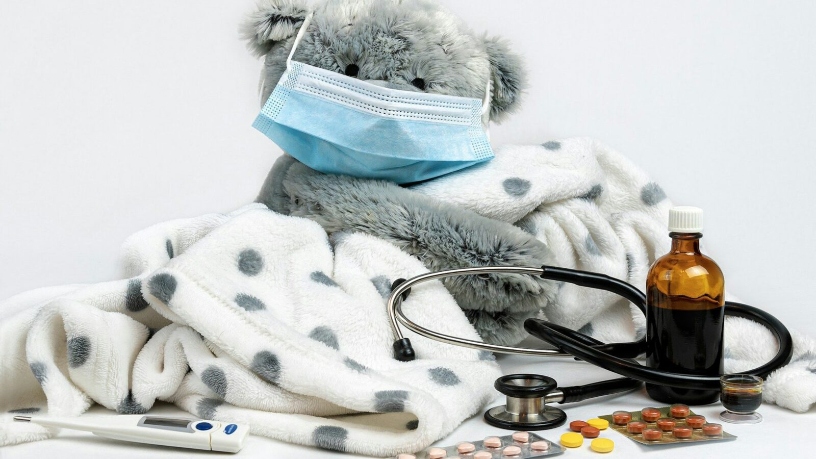 A gray teddy bear in a face mask, wrapped in a polka-dot blanket beside a stethoscope and medicine