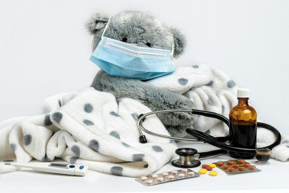 A gray teddy bear in a face mask, wrapped in a polka-dot blanket beside a stethoscope and medicine